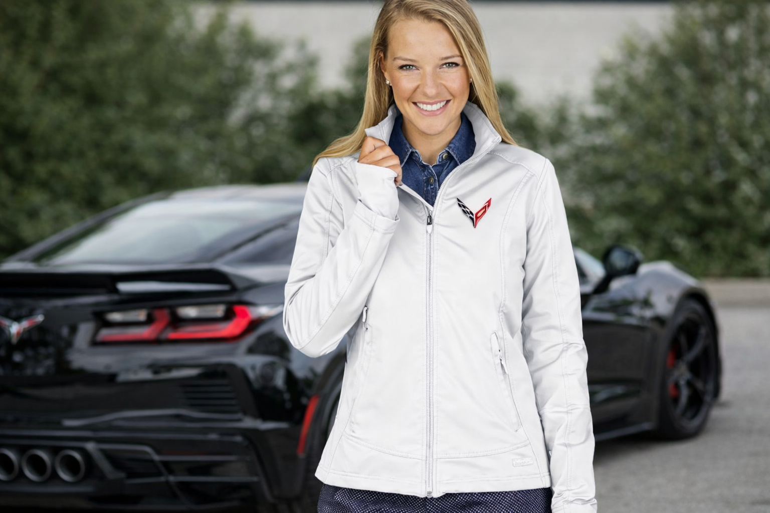 Woman wearing Corvette jacket standing in front of Corvette sports car, showcasing Corvette apparel for men and women