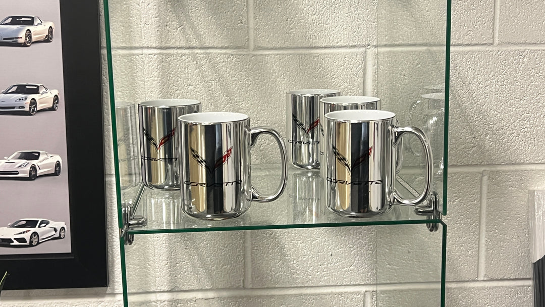 Four reflective silver ceramic mugs with a Corvette logo printed on them, displayed on a glass shelf against a white cinderblock wall.