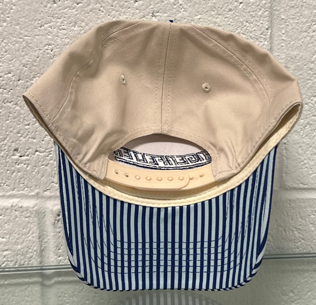 Beige cap with a blue and white striped pattern on the brim, against a textured white wall.