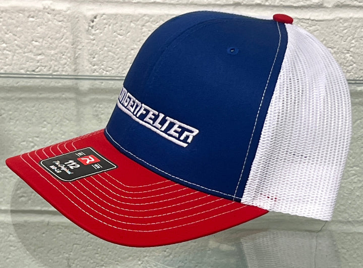 A blue, white, and red trucker hat with 'Lingenfelter' embroidered on the front panel, displayed on a glass surface against a white brick wall.