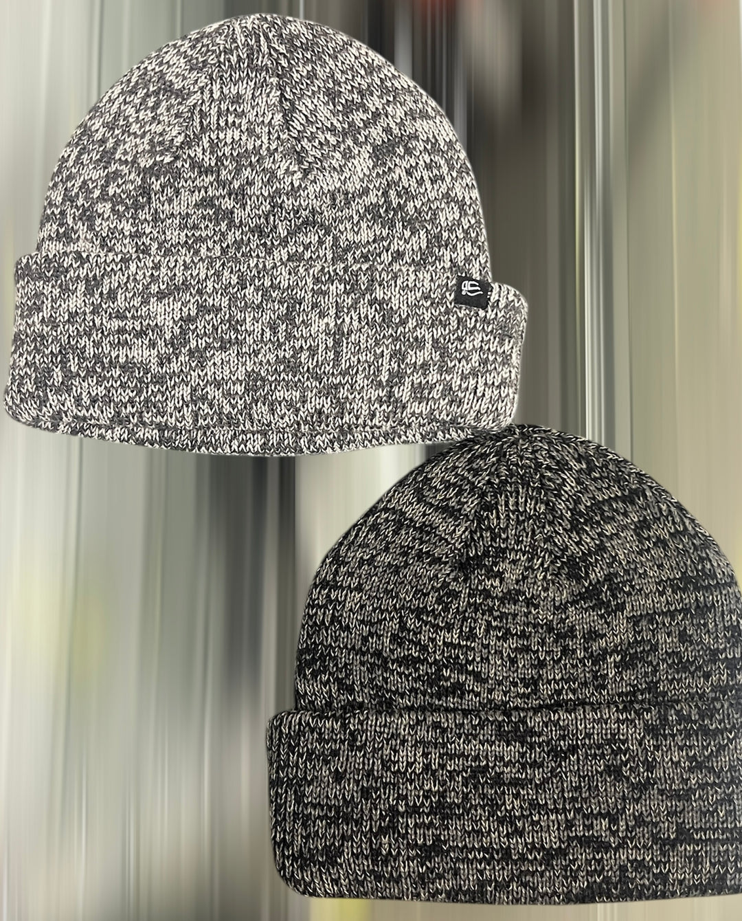 Two black and gray knit beanie hats with a brand logo on a blurred background