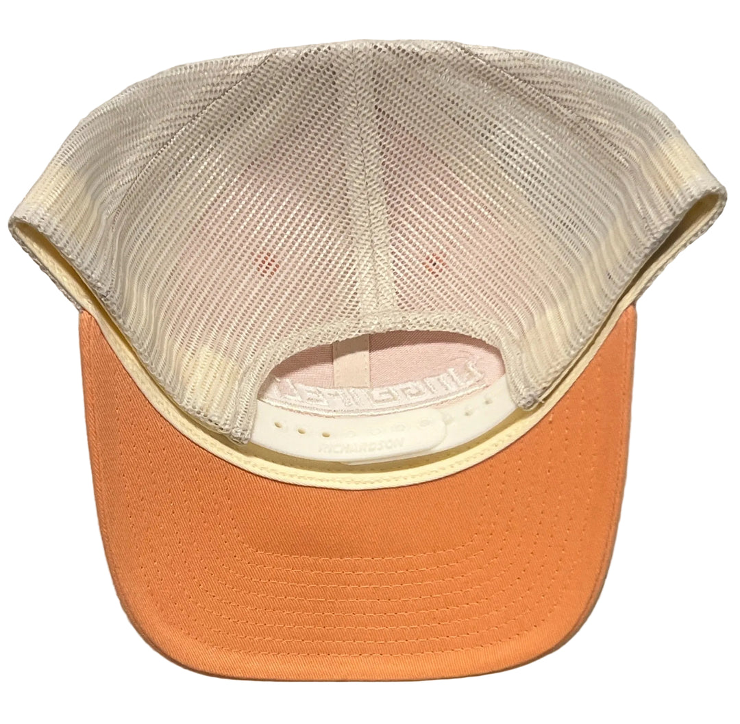 A trucker-style cap viewed from the back, featuring a peach-colored fabric brim and a cream-colored mesh back, set against a light gray textured wall.