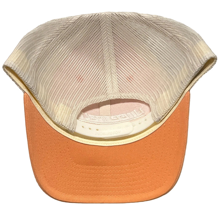A trucker-style cap viewed from the back, featuring a peach-colored fabric brim and a cream-colored mesh back, set against a light gray textured wall.