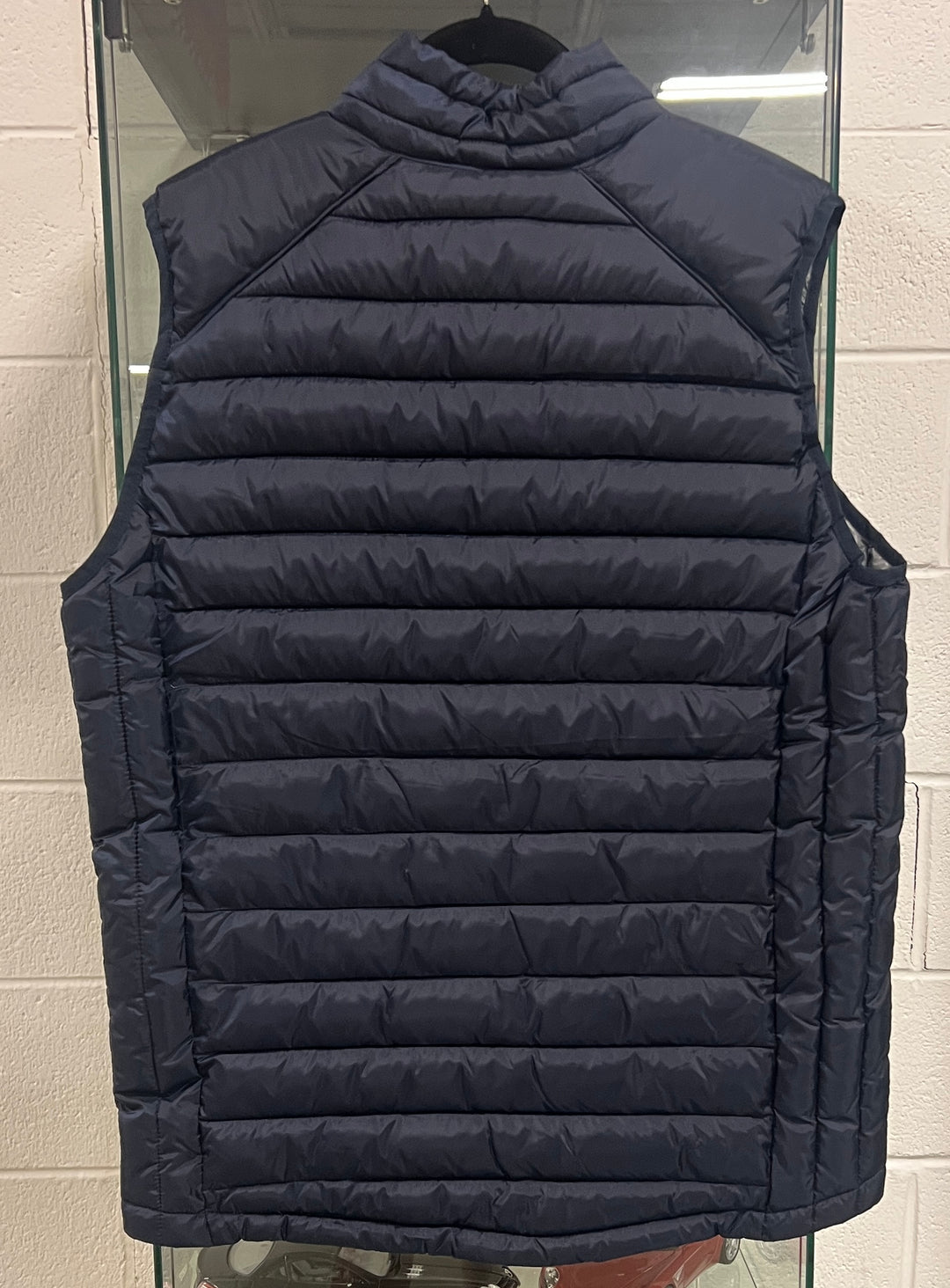 A navy blue sleeveless puffer vest with horizontal quilting, shown hanging on a black hanger against a light gray brick wall.
