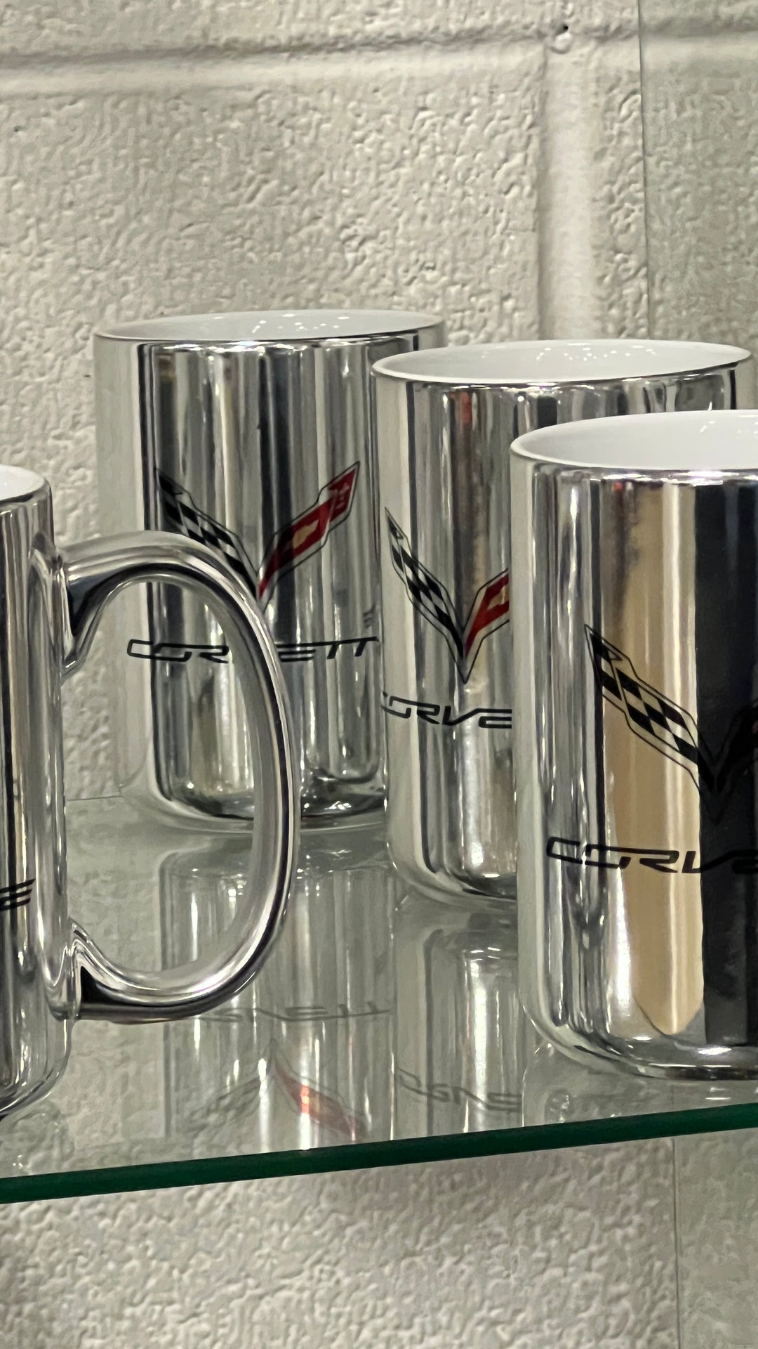 A set of shiny silver ceramic mugs with the Corvette logo printed on them, displayed on a glass shelf against a white textured wall.