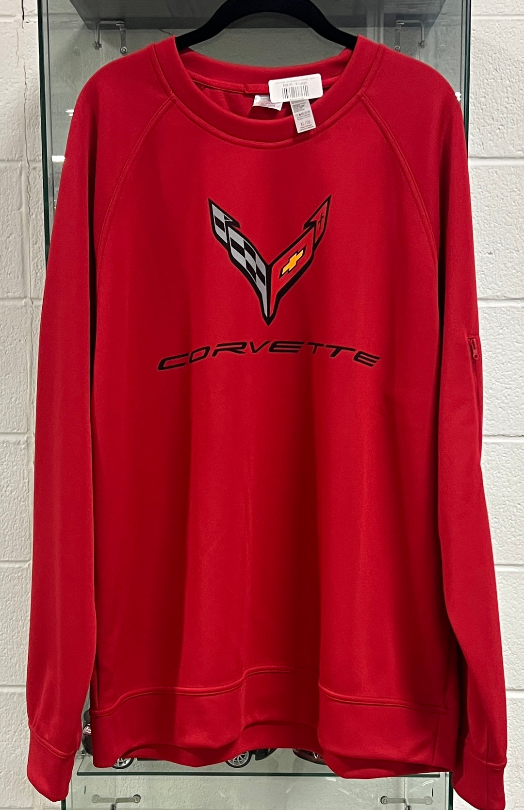 A red long-sleeve crewneck sweatshirt hanging on a black hanger against a white brick wall. The sweatshirt features a black and red Corvette logo on the chest with the word 'CORVETTE' printed in black underneath.