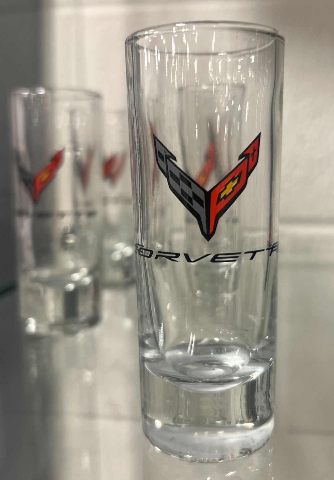 A clear glass shot glass featuring the red, black, and yellow Corvette logo and the word C8 'CORVETTE' printed on it, shown with two blurred matching glasses in the background.