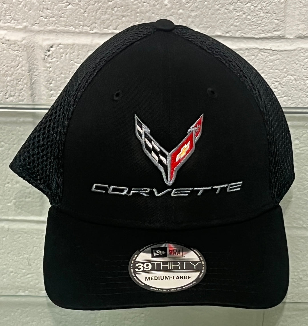 Black Corvette trucker hat with a mesh back, featuring a silver, black, and red embroidered Corvette logo on the front and a New Era 39THirty size 39 sticker on the visor, set against a white brick wall background.