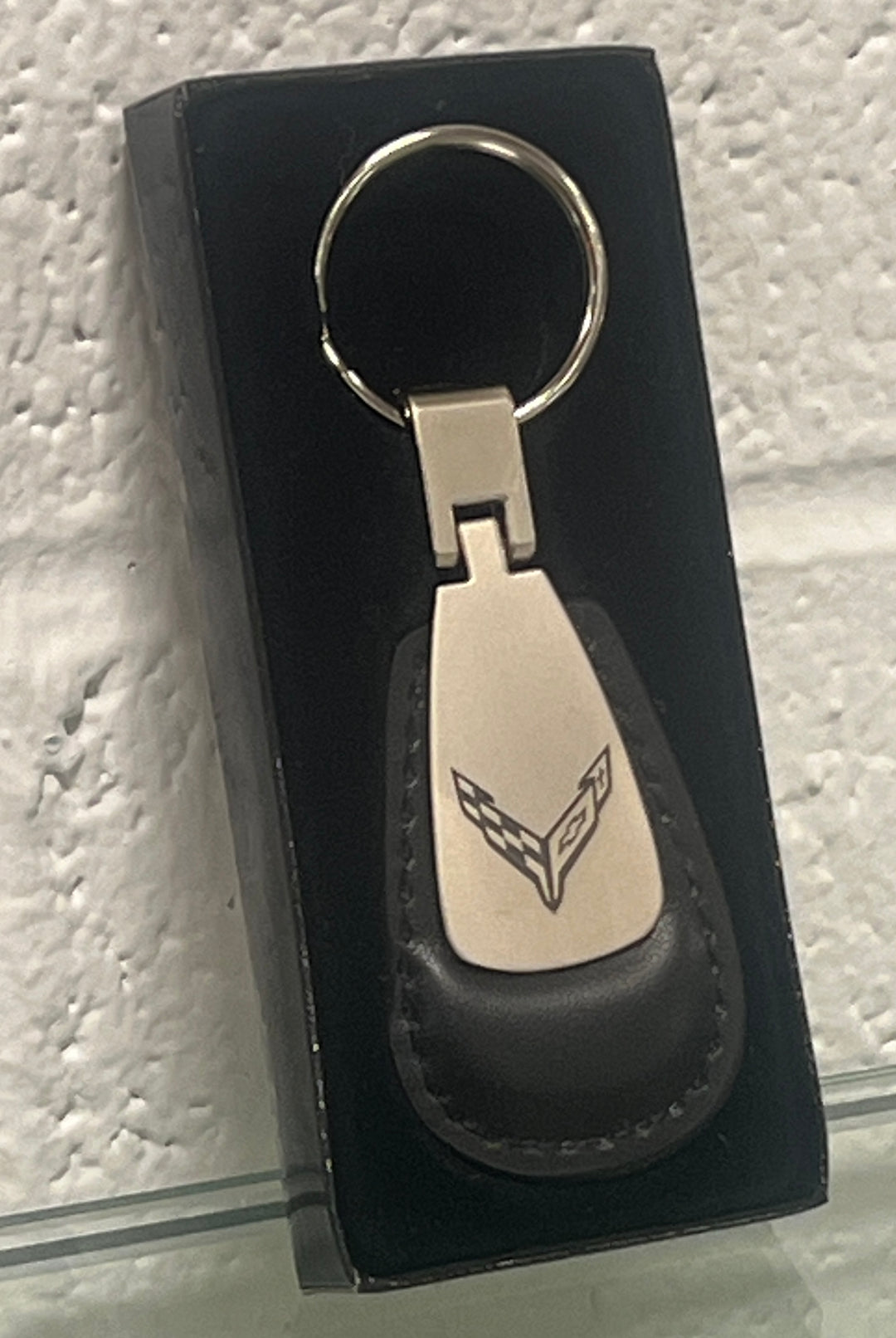A silver-tone metal keychain with a black leather tag featuring an engraved Corvette emblem, presented in a black gift box on a textured white background.