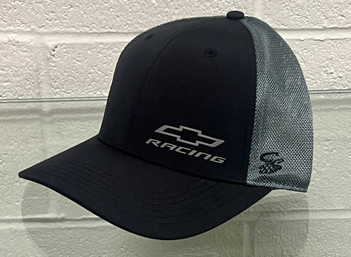 A black and gray trucker-style baseball cap with a Chevrolet Racing logo on the front, displayed against a white brick wall background.