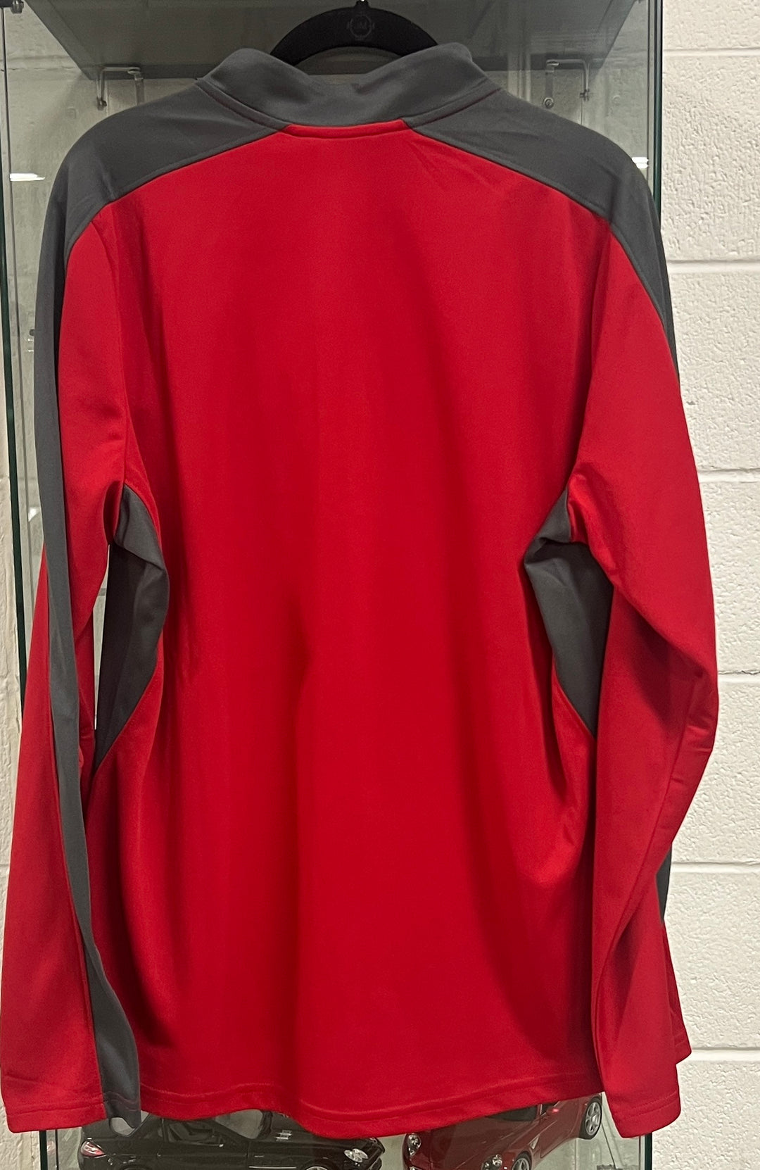 A red and gray athletic long-sleeve quarter-zip pullover jacket hanging on a black hanger against a white brick wall.
