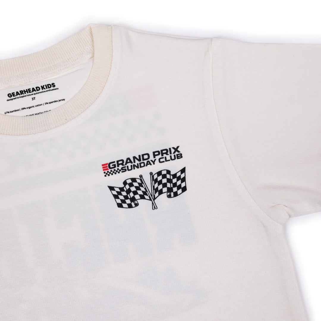 A close-up of a white short-sleeve t-shirt with a 'Grand Prix Sunday Club' graphic on the left chest, featuring a black and red logo and two checkered flags. The inner neck label shows the brand 'GEARHEAD KIDS'.