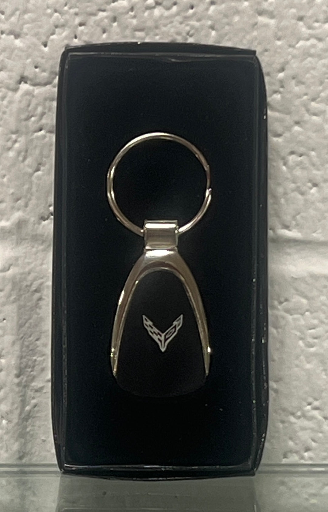 A black and silver car keychain with a winged emblem, presented in a black display box against a textured white wall.