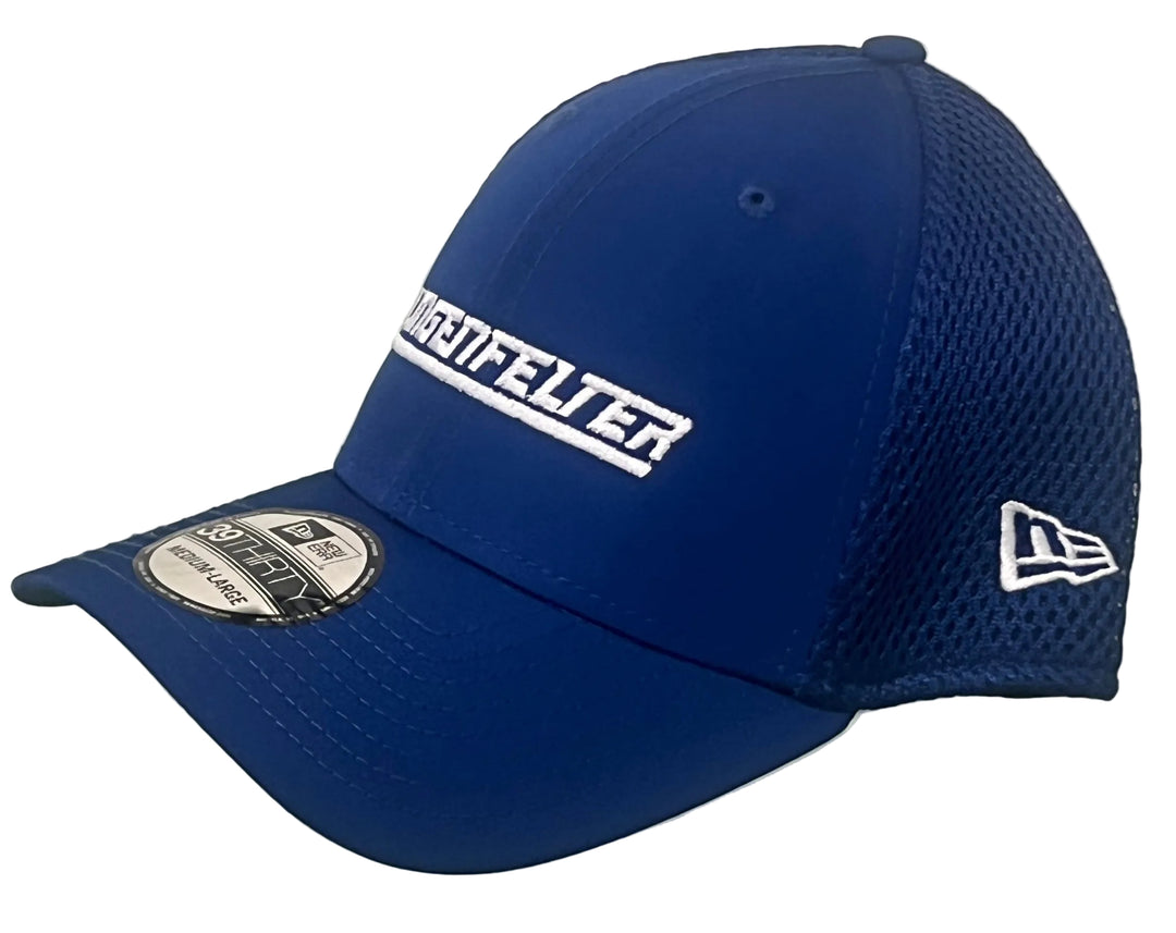 A royal blue New Era 9FORTY baseball cap with white embroidered lettering on the front and side, displayed on a clear acrylic stand against a white brick wall.