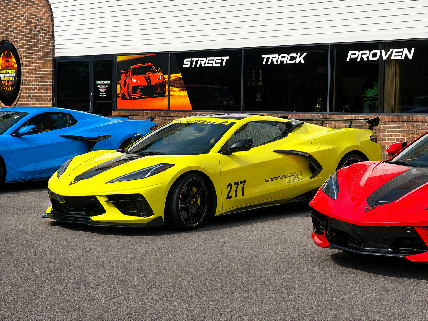 A bright yellow Corvette sports car with black racing stripes and the number 277 parked outdoors in front of a building with large windows labeled STREET, TRACK, and PROVEN. Other cars are partially visible on both sides.